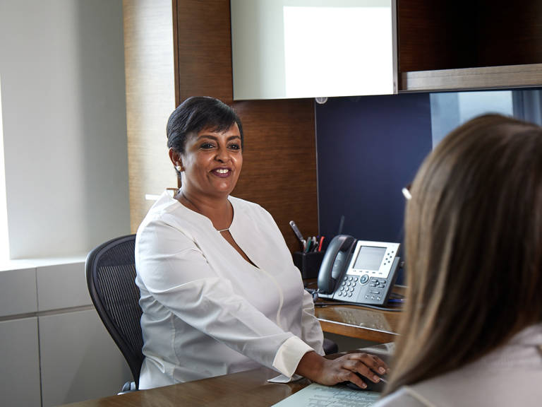 Lazard Woman Sitting at Desk Talking to Colleague Across from Her